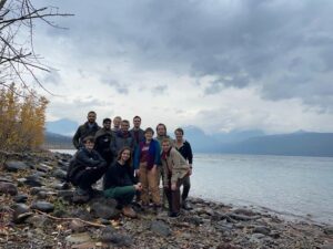 A group of scientists stand in front of a lake, with mountains in the background.