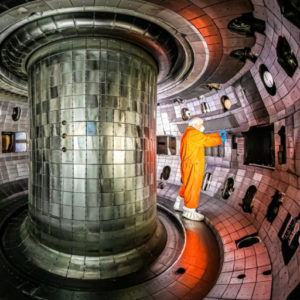 A man in orange coveralls, gloves, botties and a bonnet works inside the DIII-D vessel